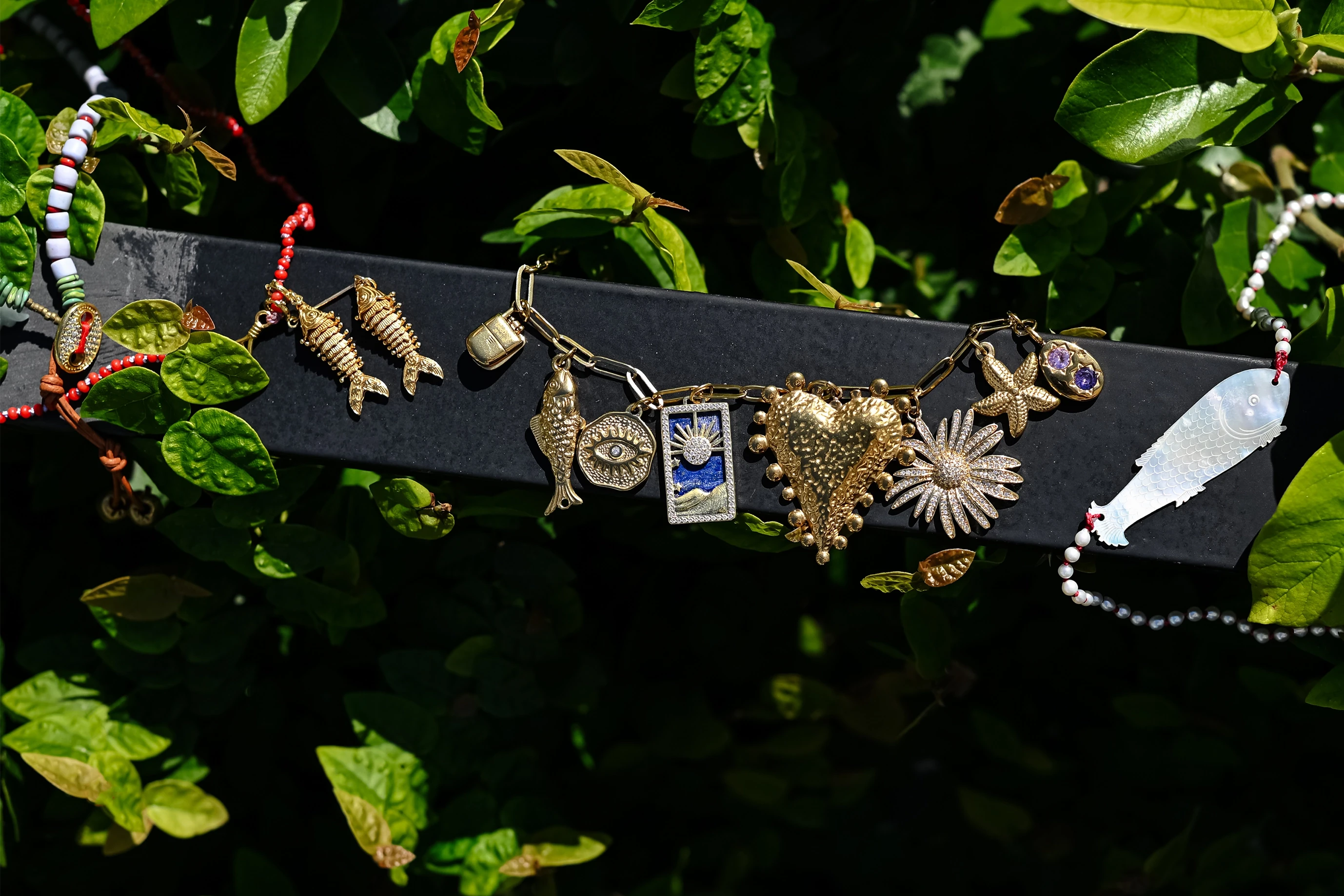 A collection of handmade jewelry charms and necklaces displayed on a black bar, set against a background of lush green leaves. The jewelry features various sea-themed motifs including a gold heart, starfish, seashell, and a small Argentina flag charm, alongside red and white beaded strands.